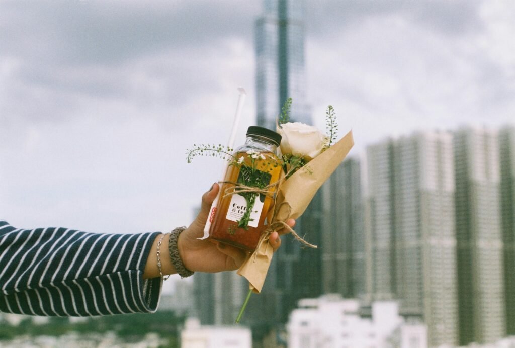 A hand holding a flower-wrapped bottle with a drinking straw, set against a city skyline on a cloudy day.