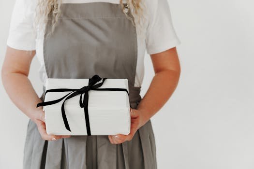 Woman in gray apron holding a white gift wrapped with black ribbon, showcasing elegance and simplicity.