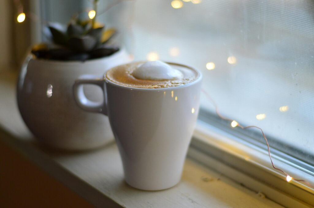 A warm latte with foam in a white cup on a windowsill next to a plant, wrapped in soft fairy lights.