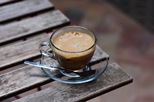 A close-up of a clear glass cup filled with frothy coffee on a rustic wooden table.