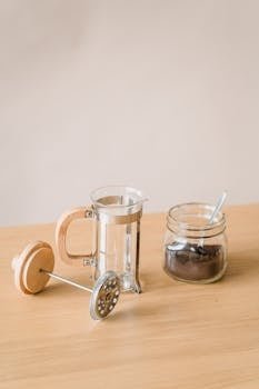 Minimalist French press and coffee jar on wooden table with soft lighting.