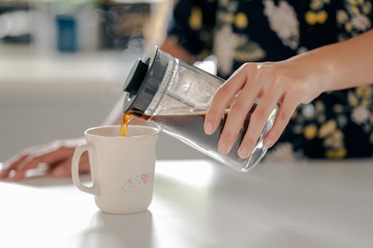 Close-up of a woman pouring coffee into a mug using a French press on a kitchen counter.