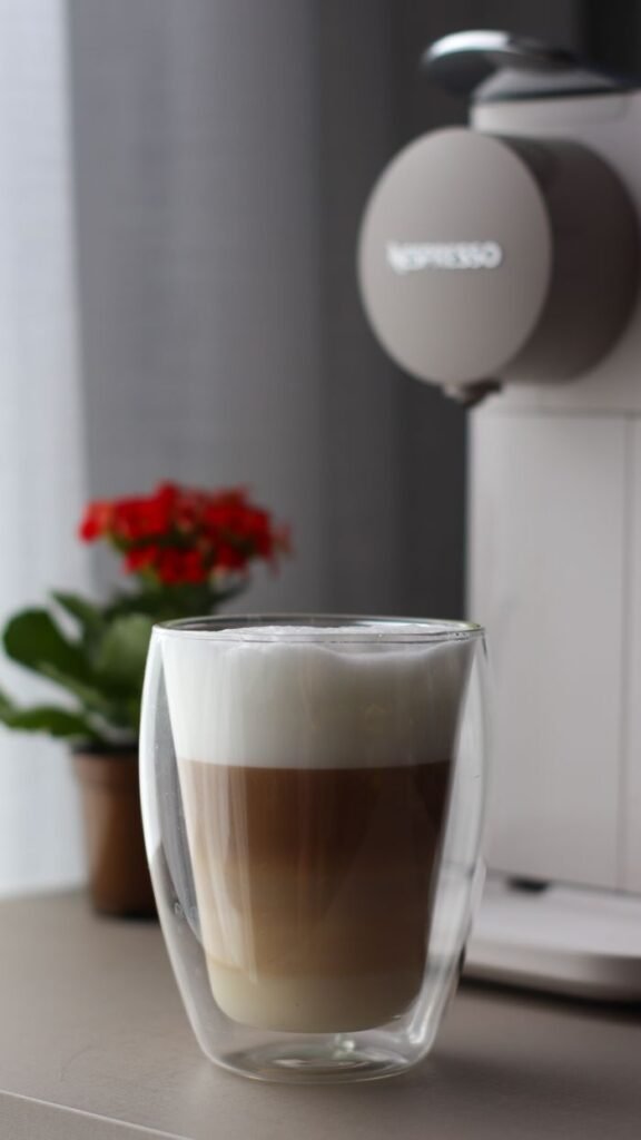 Close-up of a frothy cappuccino in a clear glass, captured indoors next to a coffee machine.