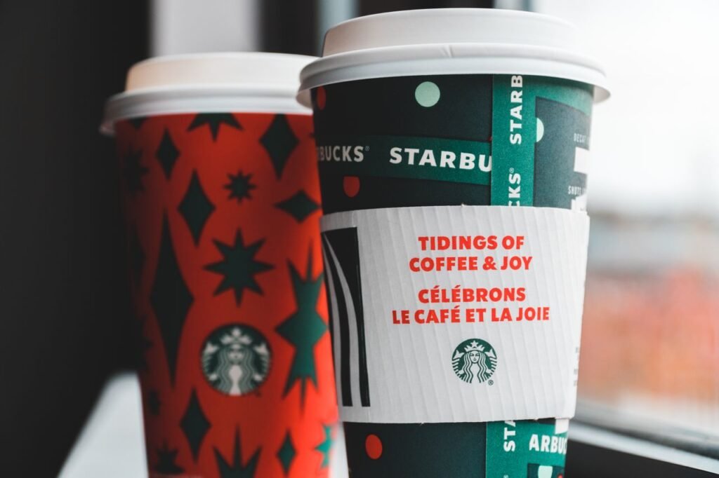 Close-up of festive Starbucks coffee cups with holiday-themed designs by a window. Starbucks Winter Drinks 2026